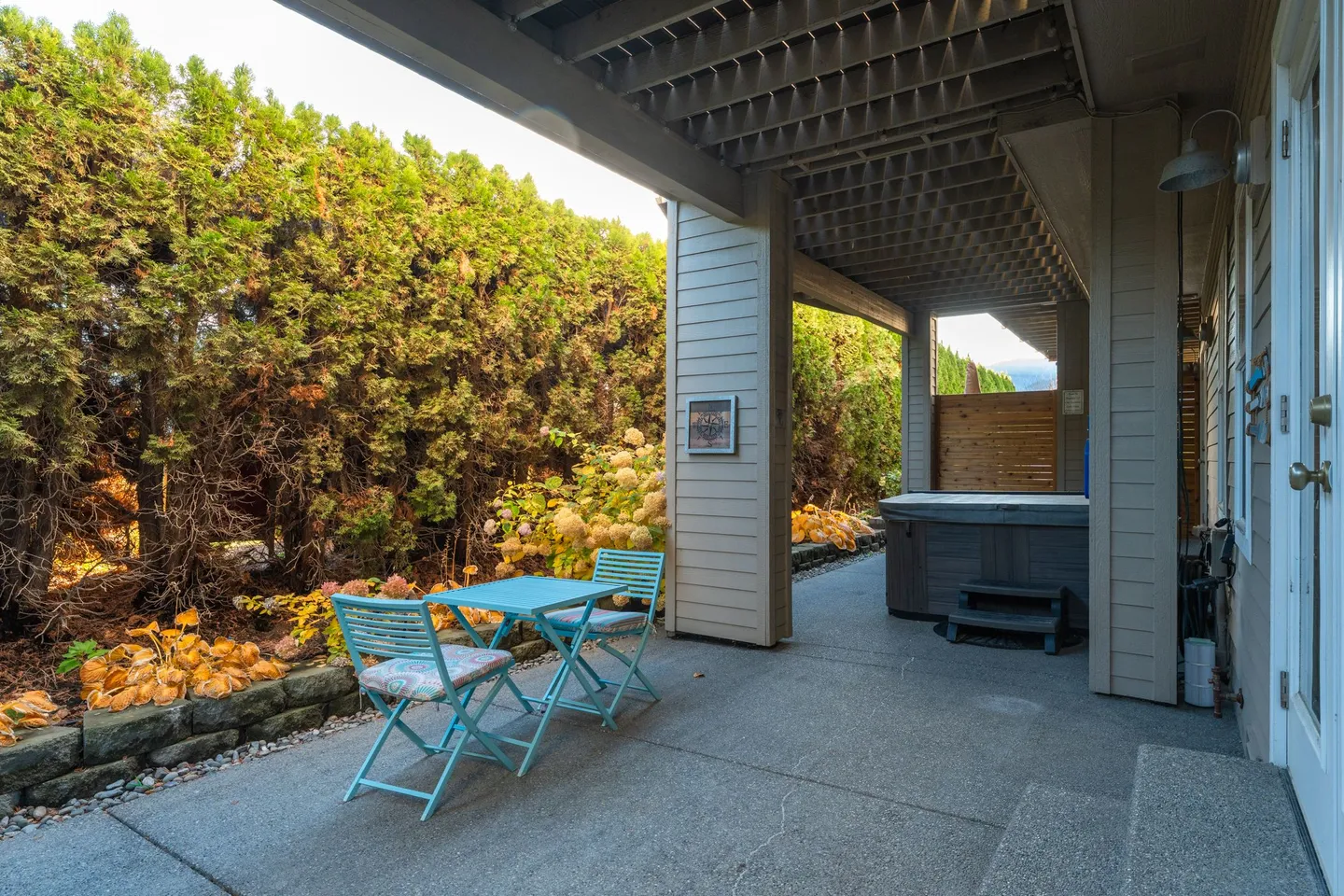 Covered concrete patio with light blue bistro set, tall evergreen hedge, and a hot tub tucked under the overhang.
