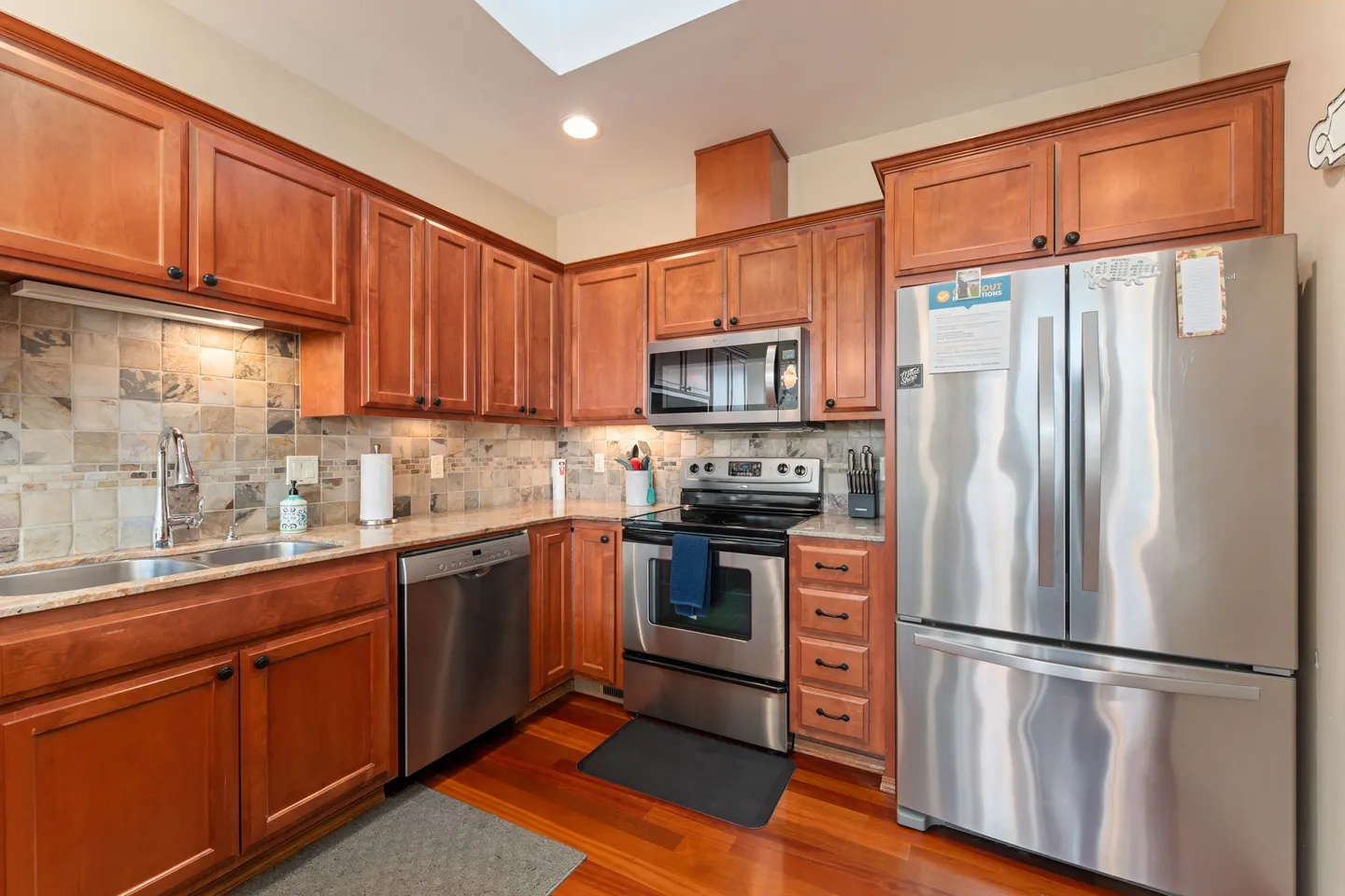 Kitchen with warm wood cabinetry, stainless steel appliances, stone tile backsplash, and wood flooring.