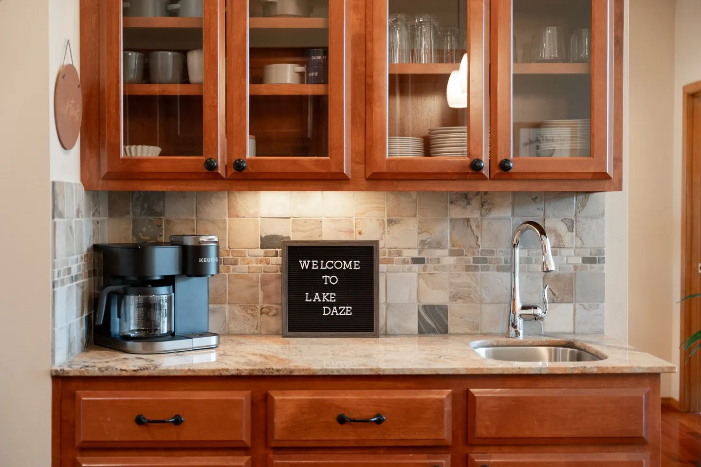 Wet bar area featuring warm wood cabinetry, a tiled backsplash, prep sink, coffee maker, and a letter board reading 'Welcome to Lake Daze'.