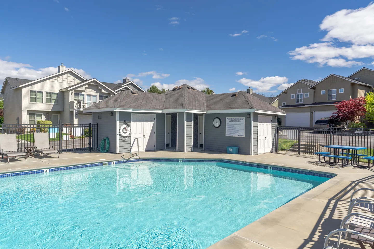 Outdoor swimming pool with clear turquoise water surrounded by a concrete deck, fence, small clubhouse building, and residential townhomes under a blue sky.