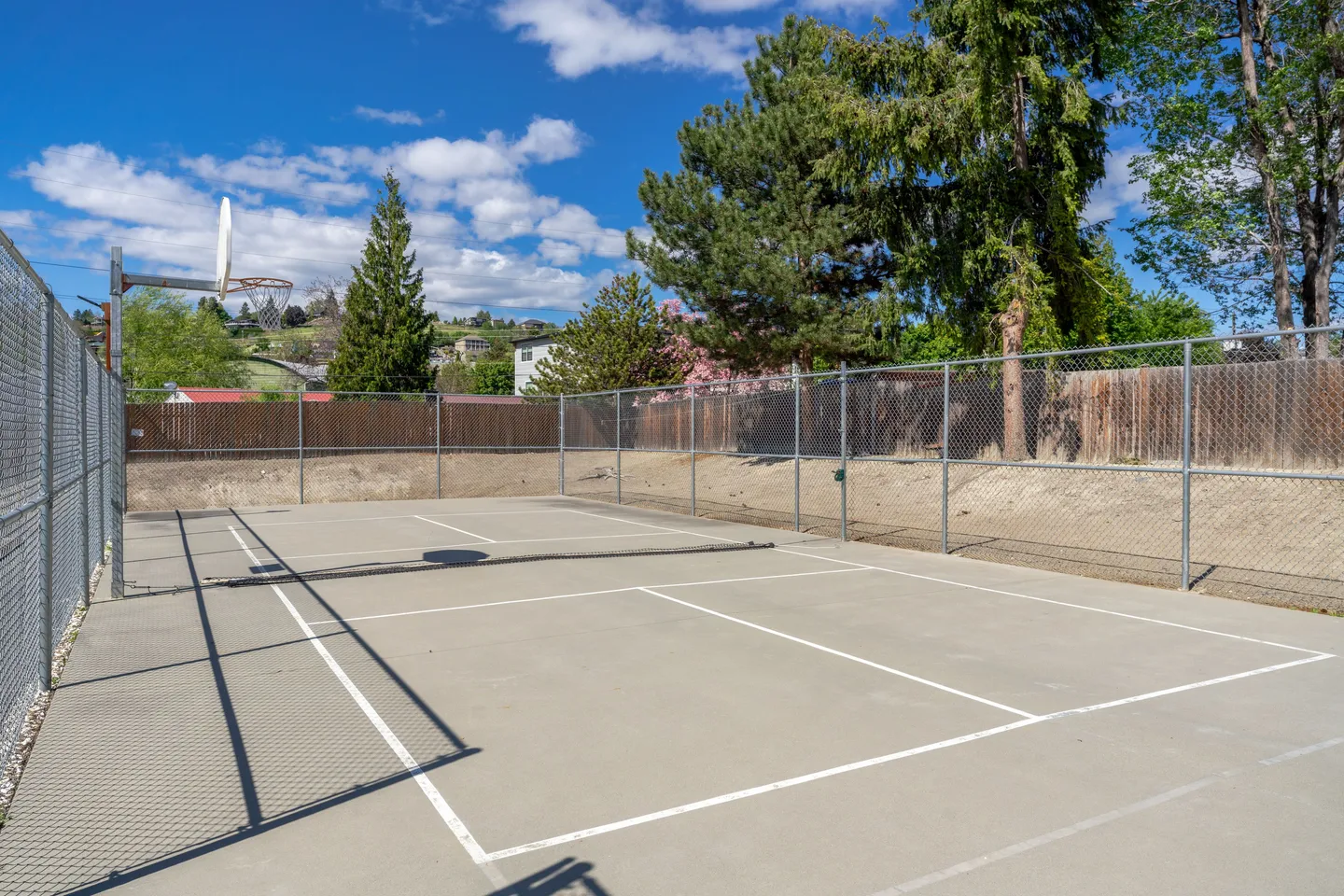 An outdoor fenced multi-use sport court with a low net, basketball hoop, and painted court lines under a blue sky.