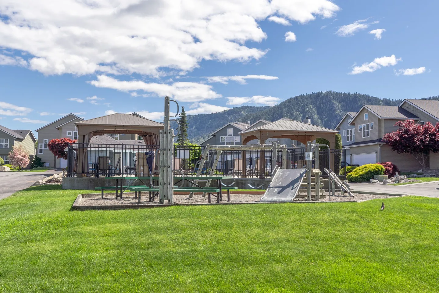Outdoor community playground and fenced pool area with gazebos in front of residential townhomes and forested mountains.