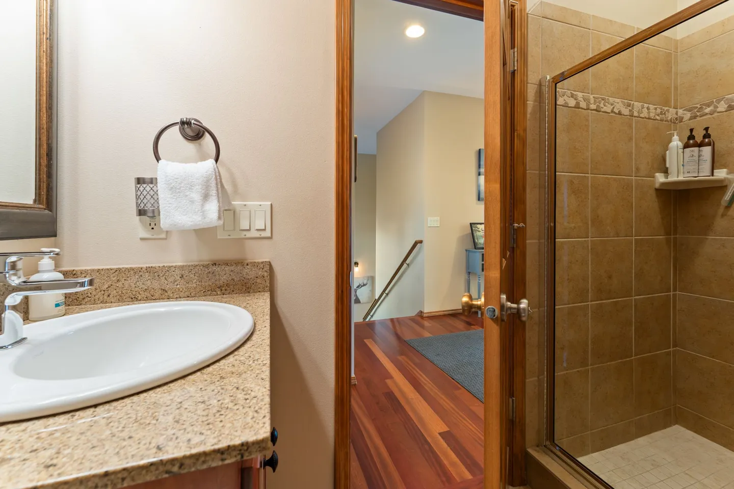 Bathroom vanity and walk-in tiled shower with an open doorway revealing a hardwood floor and staircase.
