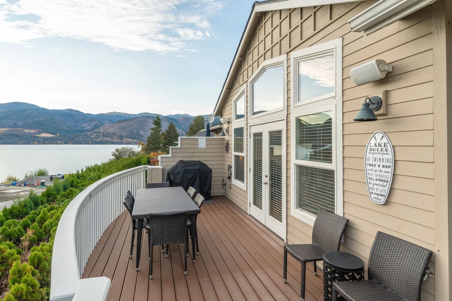 Curved outdoor deck with a dining table, chairs, barbecue grill, and panoramic views of a lake and mountains.