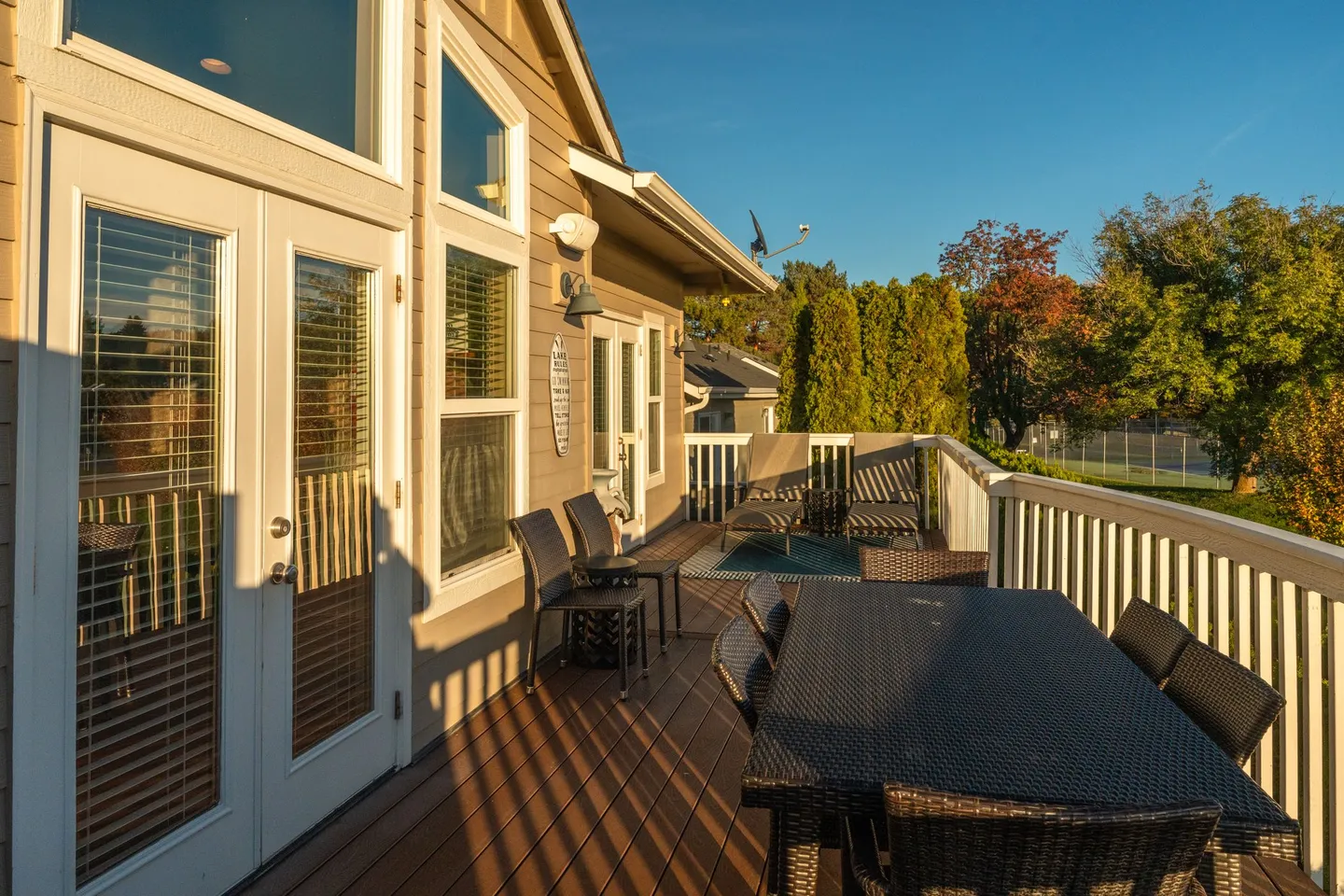 Outdoor wooden deck with outdoor dining table, chairs, lounge seating, and white railings under a clear blue sky.