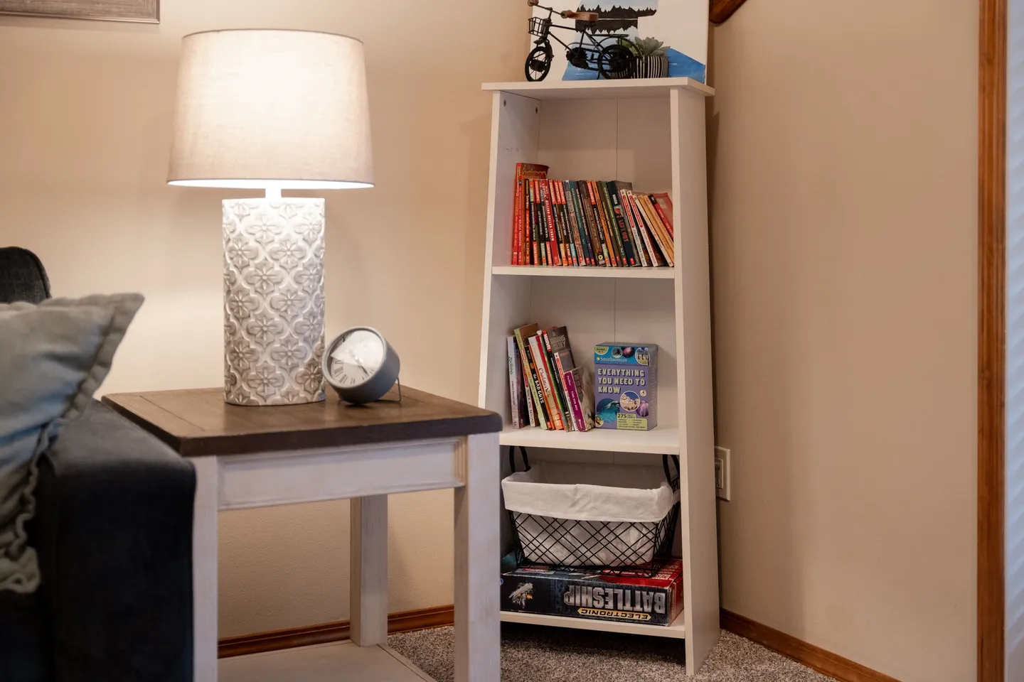 A corner reading nook featuring a white side table with a patterned lamp, an alarm clock, and a slim white bookshelf holding books and games.
