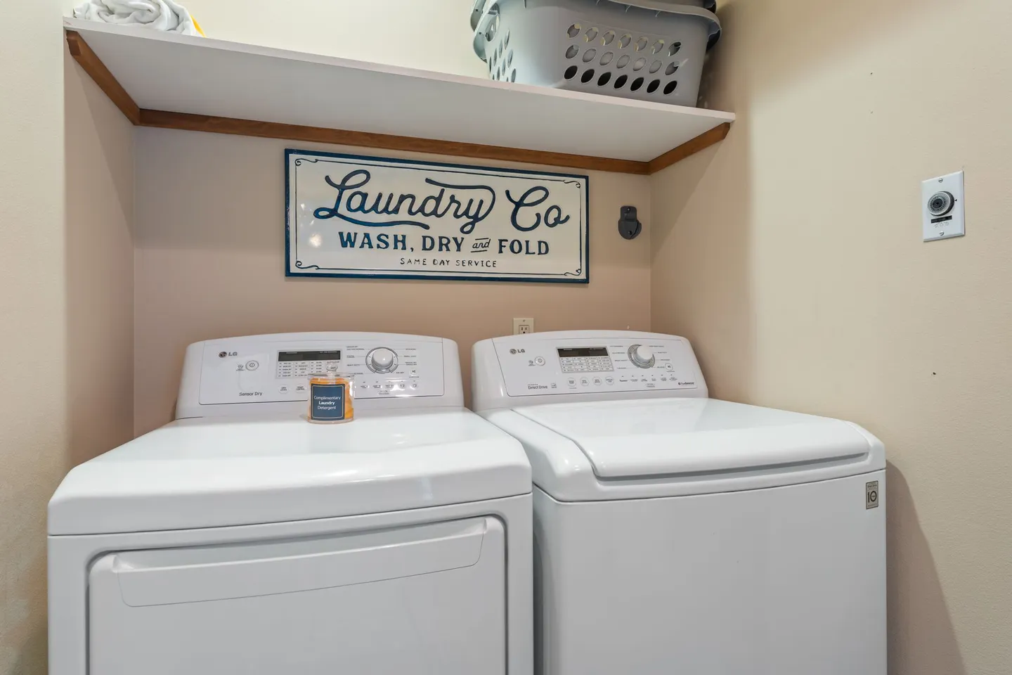 A white front-load dryer and top-load washer side by side under a wall-mounted shelf with a decorative laundry sign.