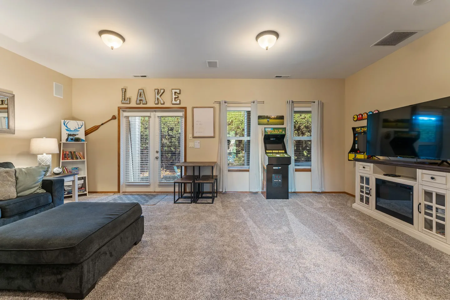 Carpeted game and living room area with a large TV on an electric fireplace console, arcade machines, small dining table, and glass patio doors.