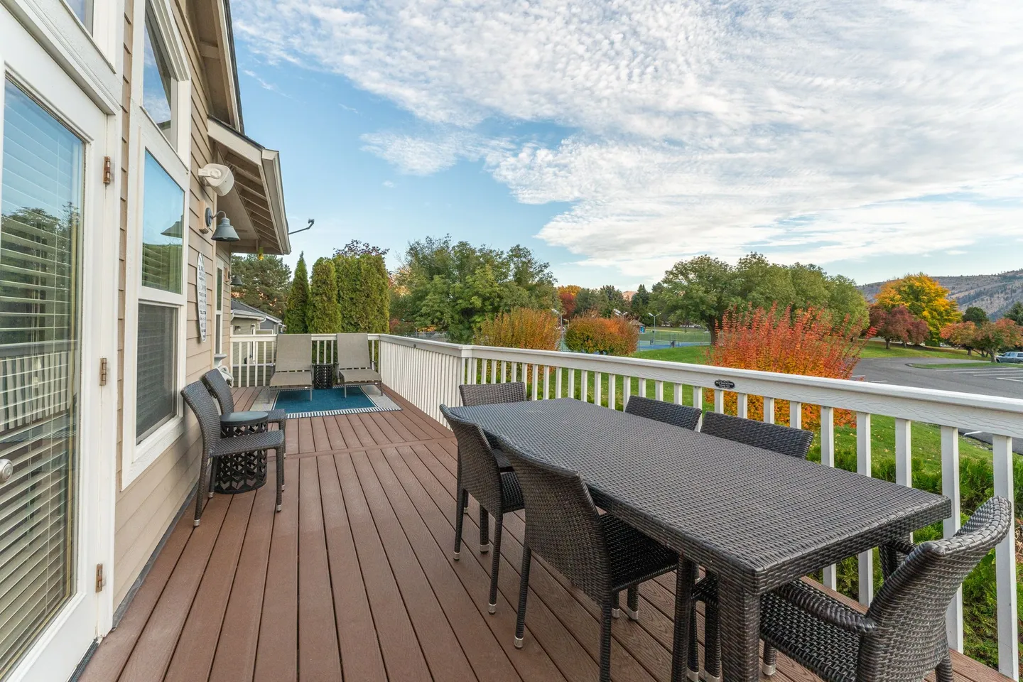 Outdoor wooden deck with a large dining table, chairs, lounge chairs, and a white railing overlooking trees and hills.