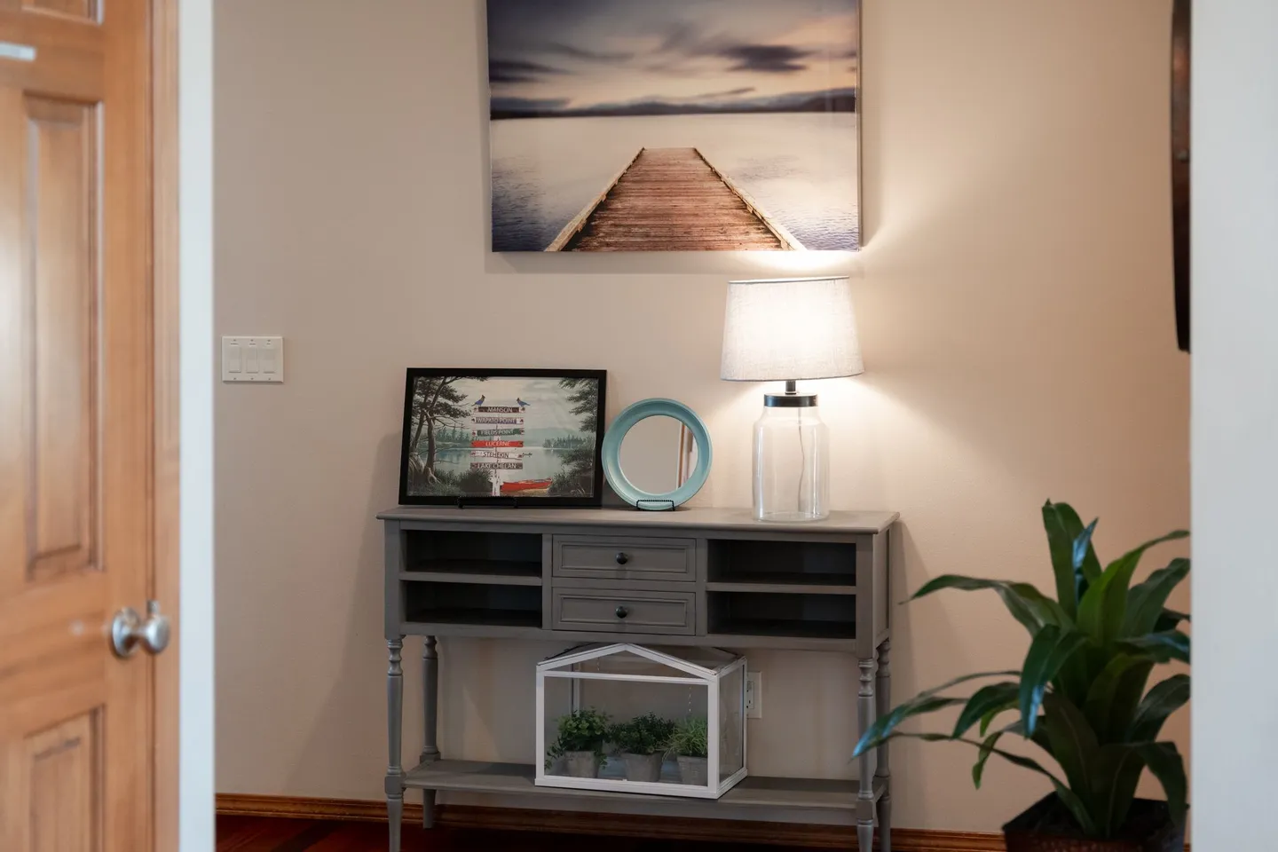 A decorated gray wooden console table set against a beige wall with a lamp and framed artwork.
