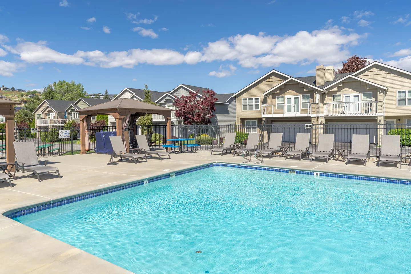 Outdoor swimming pool surrounded by a concrete patio, lounge chairs, a gazebo, and adjacent residential buildings under a blue sky.
