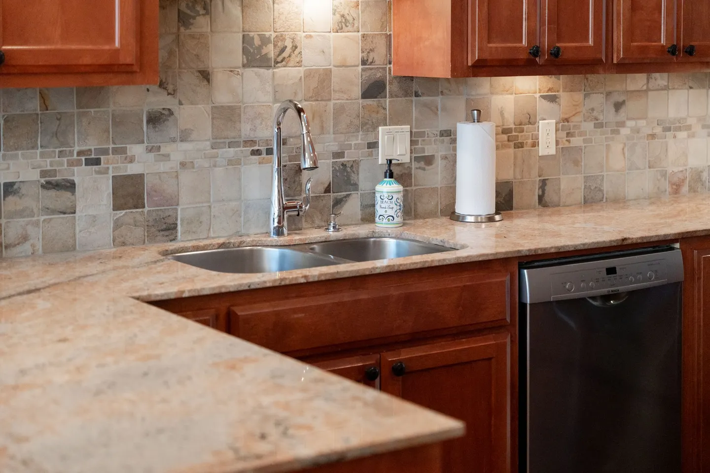 Close-up of a kitchen sink area with speckled countertops, wooden cabinets, and a tile backsplash.