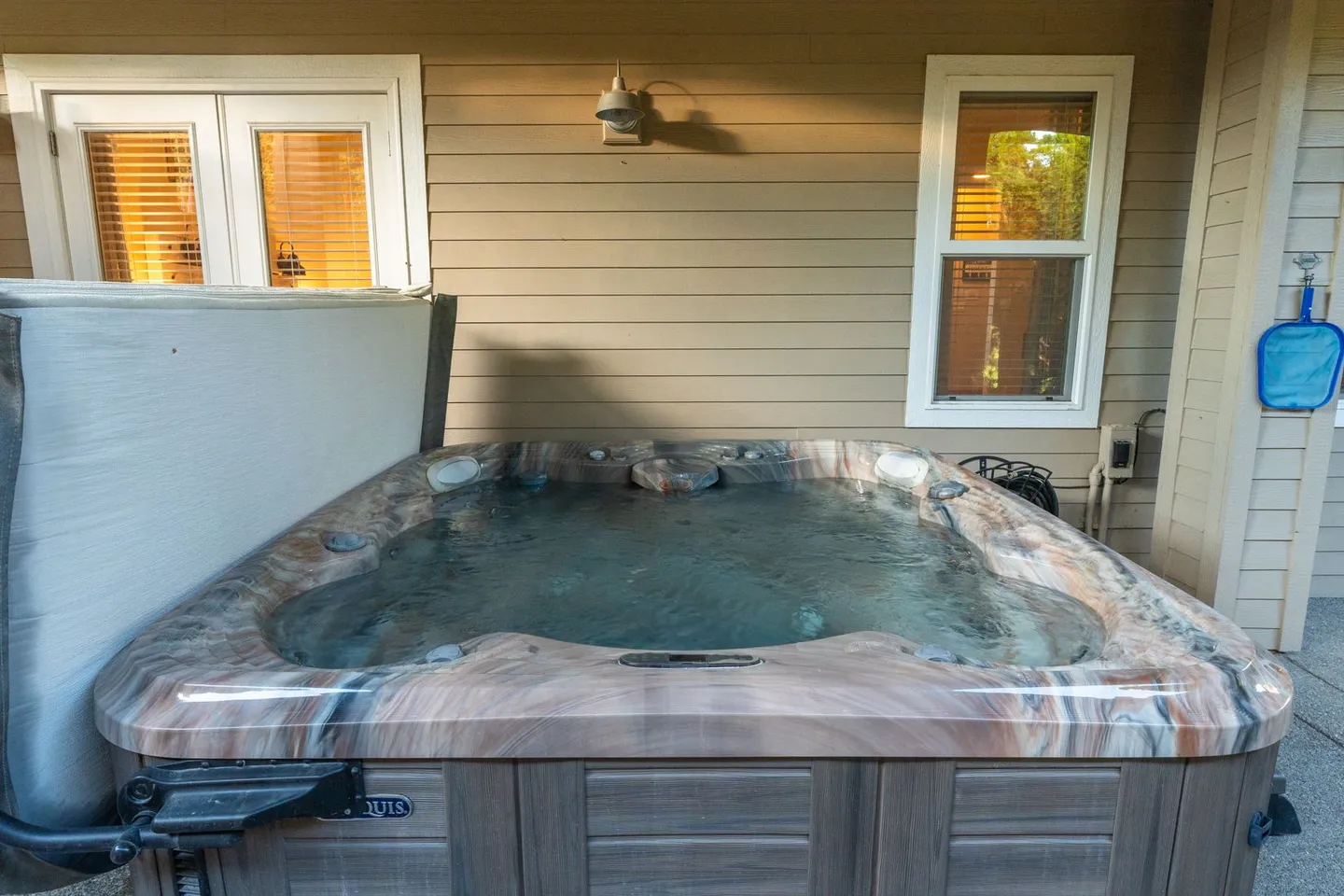 Outdoor hot tub filled with water positioned against the siding of a house with windows and a light fixture.