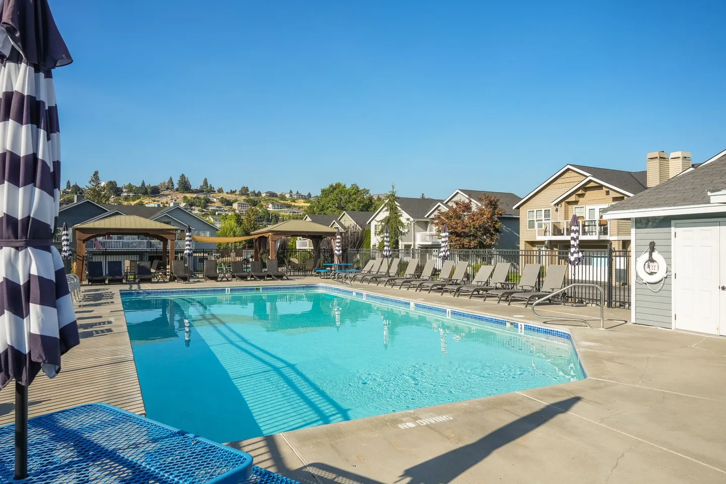 Outdoor swimming pool surrounded by concrete decking, lounge chairs, and residential buildings under a clear blue sky.