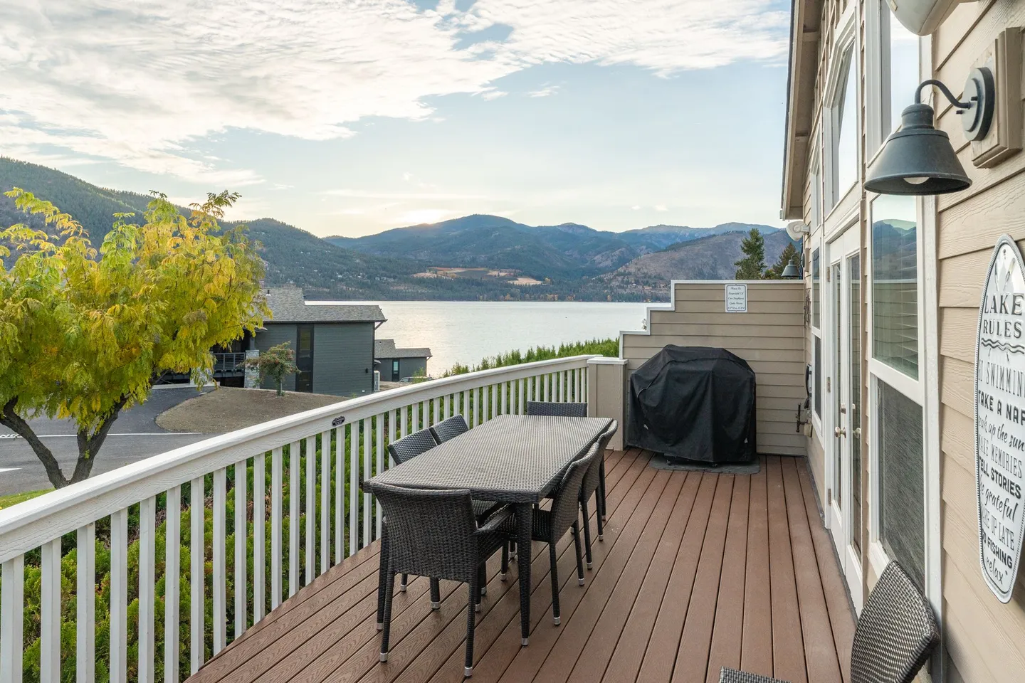 Outdoor deck with a dining table, chairs, grill under a cover, and panoramic views of a lake and mountains.