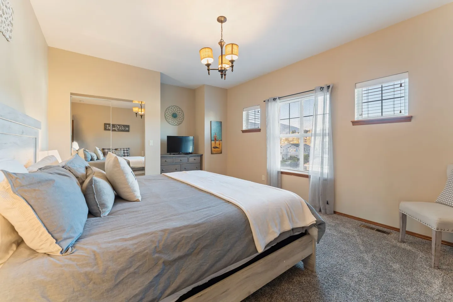 Bedroom with a made bed, carpeted floors, light beige walls, a television on a dresser, and multiple windows.