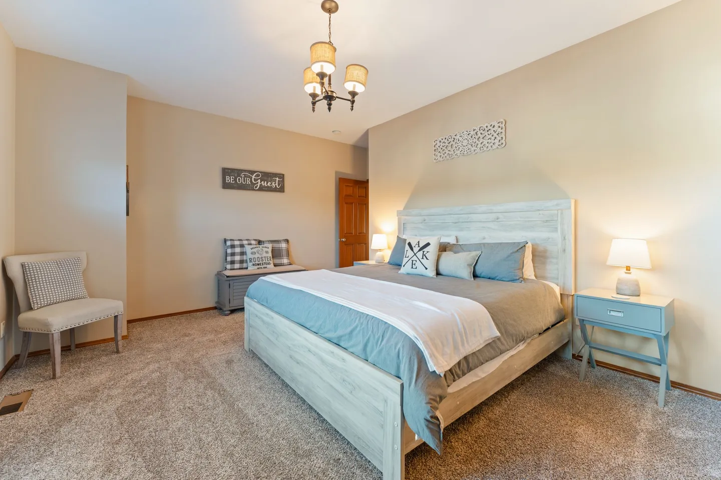 Bedroom with a wooden bed, nightstands with lamps, a bench, an accent chair, and carpeted flooring.