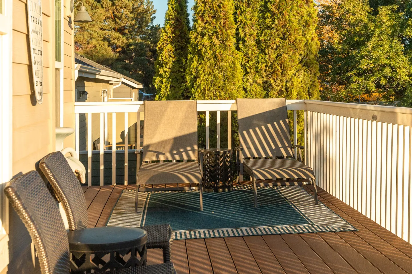 Outdoor wooden deck with lounge chairs, armchairs, and an outdoor rug surrounded by white railings and trees.