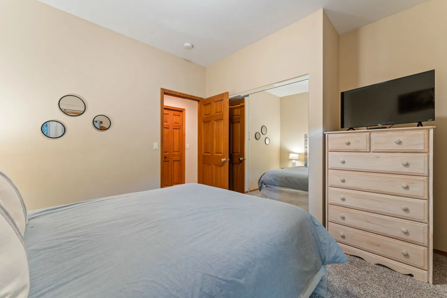 Bedroom featuring a made bed with a light blue comforter, mirrored sliding closet doors, and a light wood dresser with a flat-screen television.
