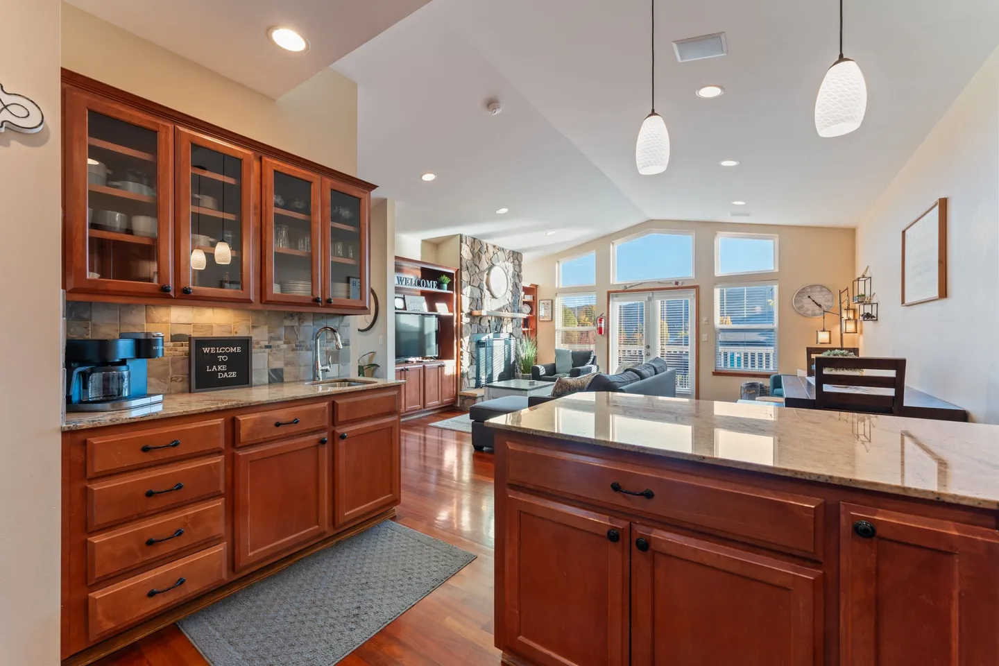 Open-concept kitchen area looking toward the living room with stone fireplace, dining table, and large windows.
