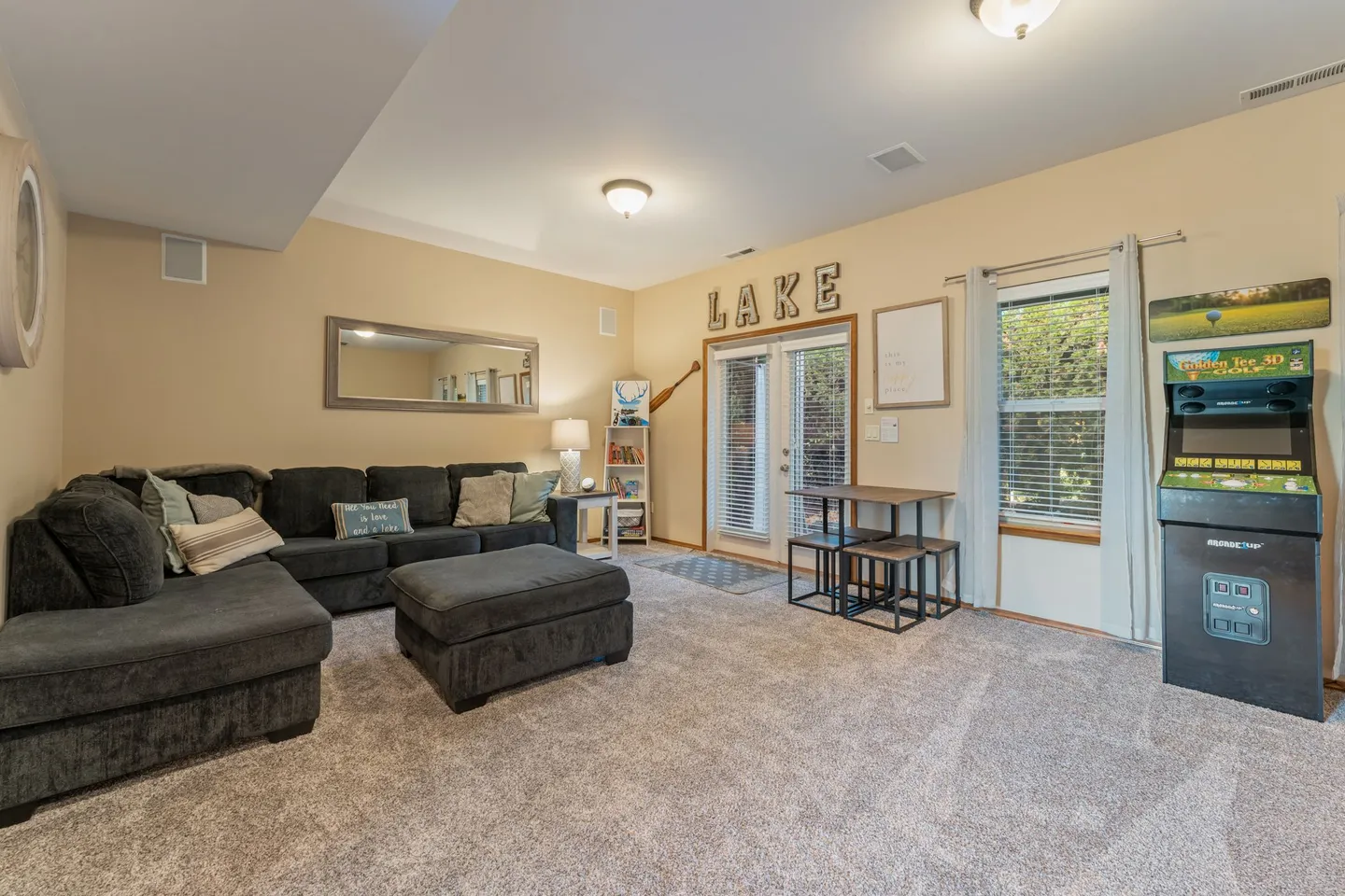 Living area with a dark sectional couch, matching ottoman, carpet flooring, a small table set, and an arcade game cabinet.