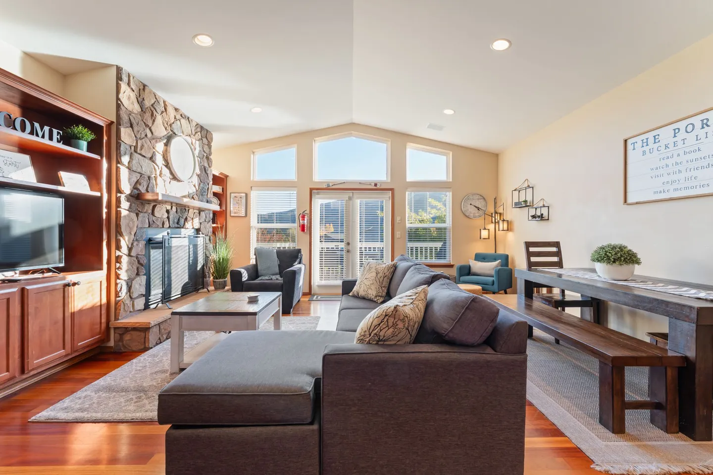 Living room with vaulted ceiling, sectional couch, stone fireplace, and adjacent dining area with a long wooden bench table.