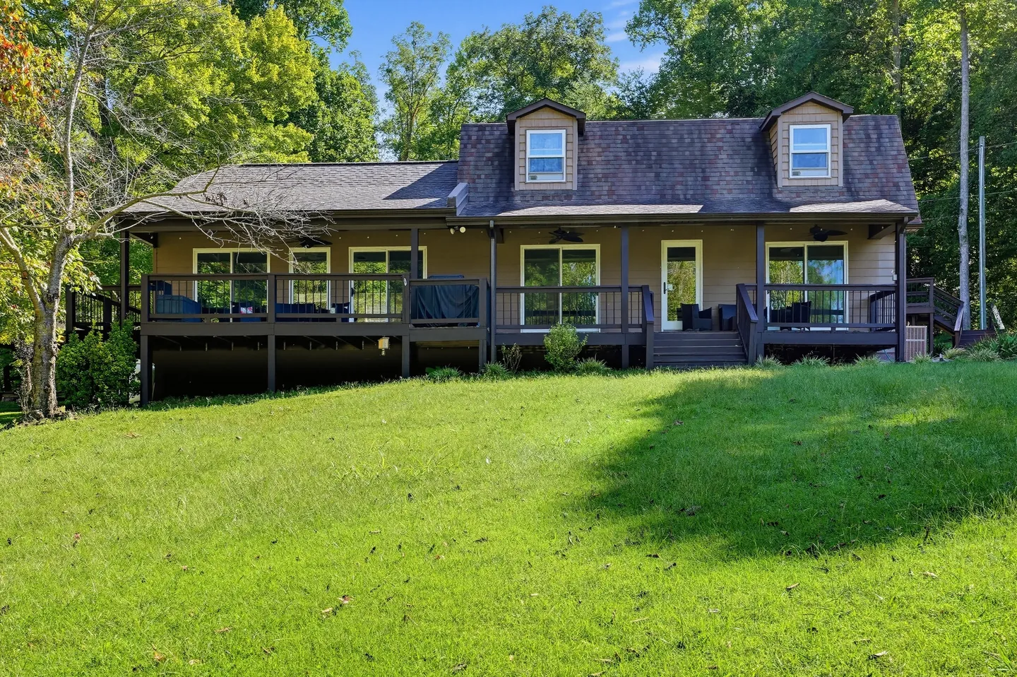 Exterior view of the back of a house with an elevated wooden deck overlooking a grassy sloped lawn bordered by trees.