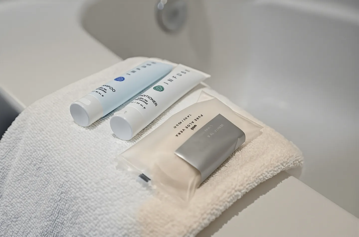 Close-up of travel-sized toiletries, including shampoo, conditioner, and bar soap, resting on a folded towel along the edge of a white bathtub.