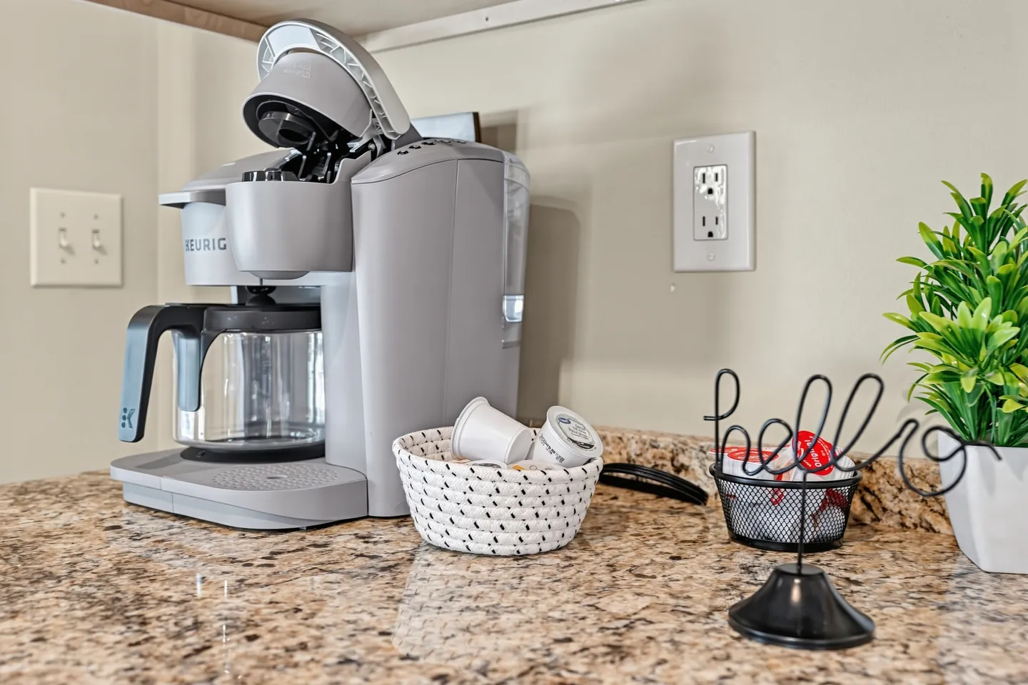 Close-up of a gray Keurig coffee maker with a carafe on a granite countertop next to baskets of coffee pods and a decorative hello sign.