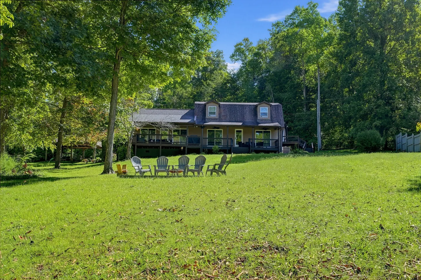 Exterior view of a multi-level house with a deck overlooking a large grassy yard featuring a fire pit and chairs surrounded by mature trees.