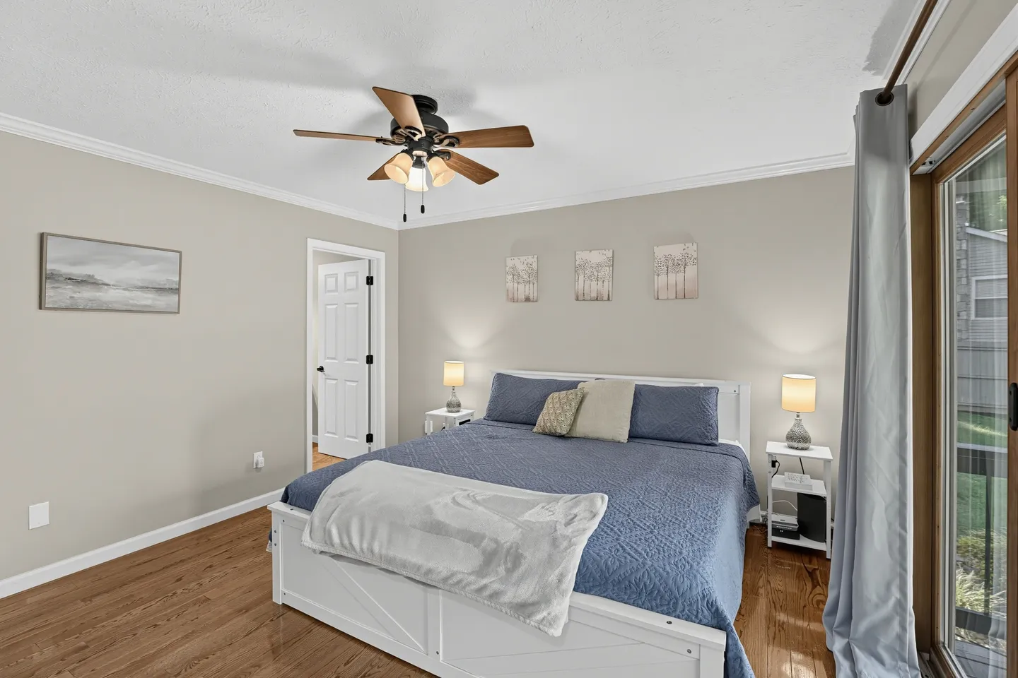 Bedroom with a white wooden bed frame, blue bedding, hardwood-style floors, a ceiling fan, and sliding glass doors.