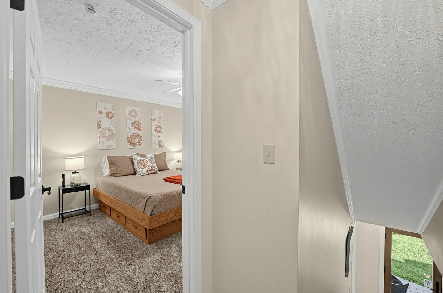 A view through an open doorway into a carpeted bedroom with a wooden bed frame, nightstand, and wall art, beside an angled stairwell.