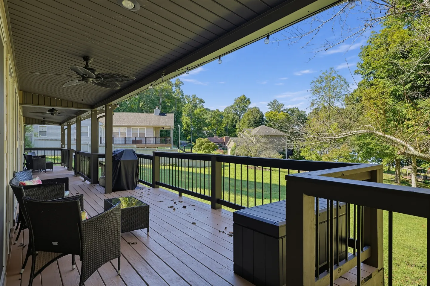 A covered back deck with outdoor chairs, a small table, a grill, and views of a green lawn and trees.