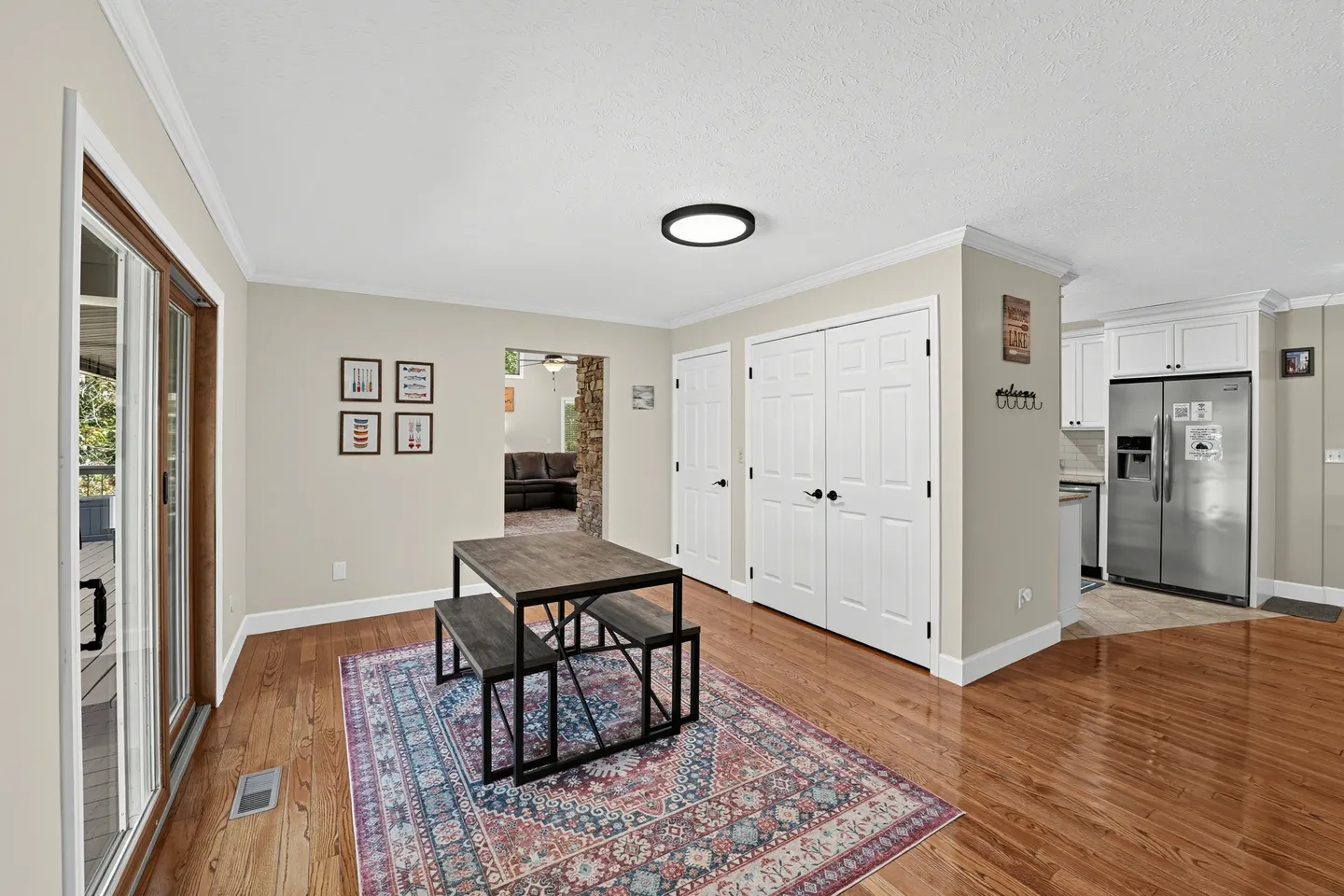 Dining area with a wooden table and benches on a patterned rug, adjacent to sliding glass doors and kitchen.