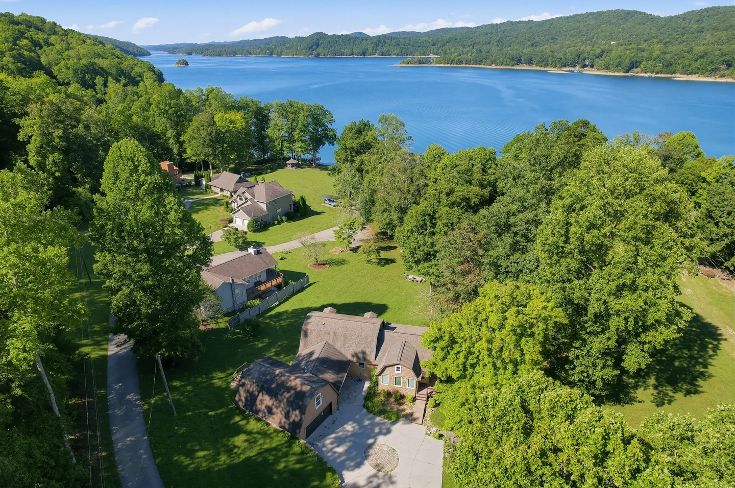 Aerial view of a lakeside home surrounded by lush green trees, overlooking a large blue body of water and forested hills.