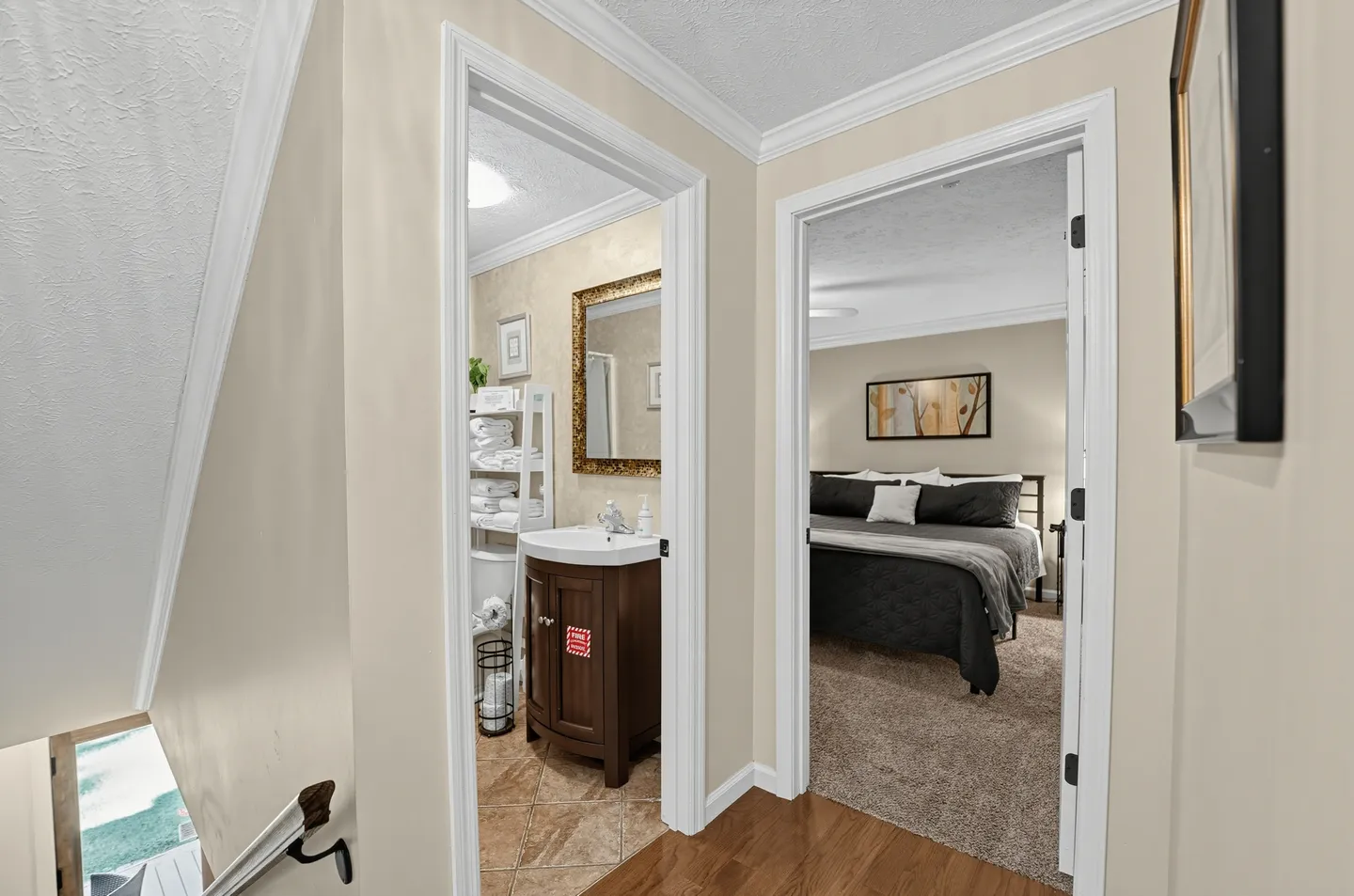 Hallway view looking into a bathroom with a dark wood vanity and an adjacent carpeted bedroom with a made bed.