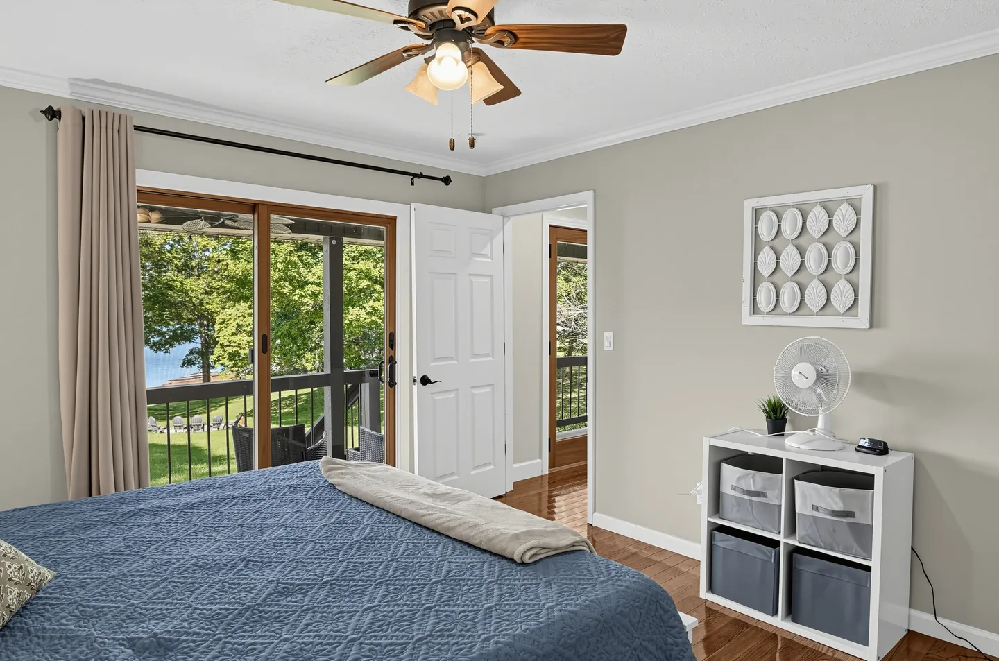 Bedroom with blue bedding, a ceiling fan, and sliding glass doors opening to an outdoor deck overlooking trees and water.