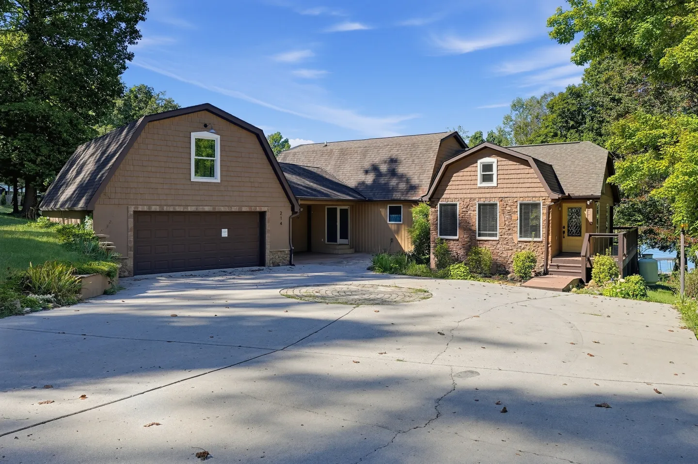 Exterior view of a multi-level home with gambrel rooflines, an attached garage, stone accents, and a large paved driveway under a blue sky.