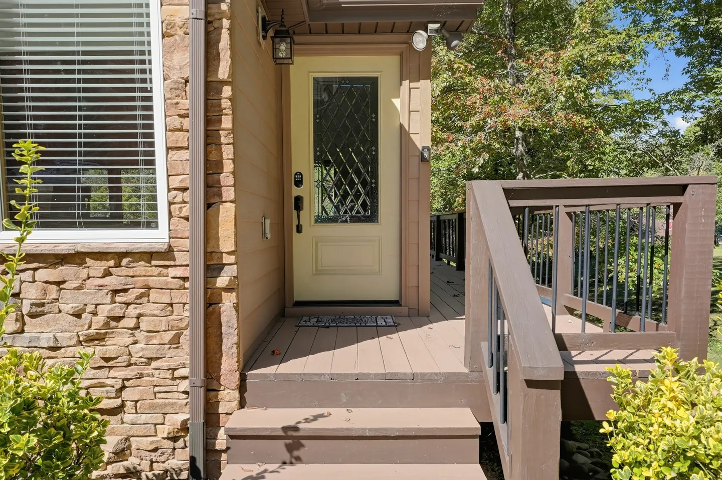 Front entryway with wooden steps leading to a yellow front door with a decorative glass panel, flanked by stone veneer siding and a deck railing.