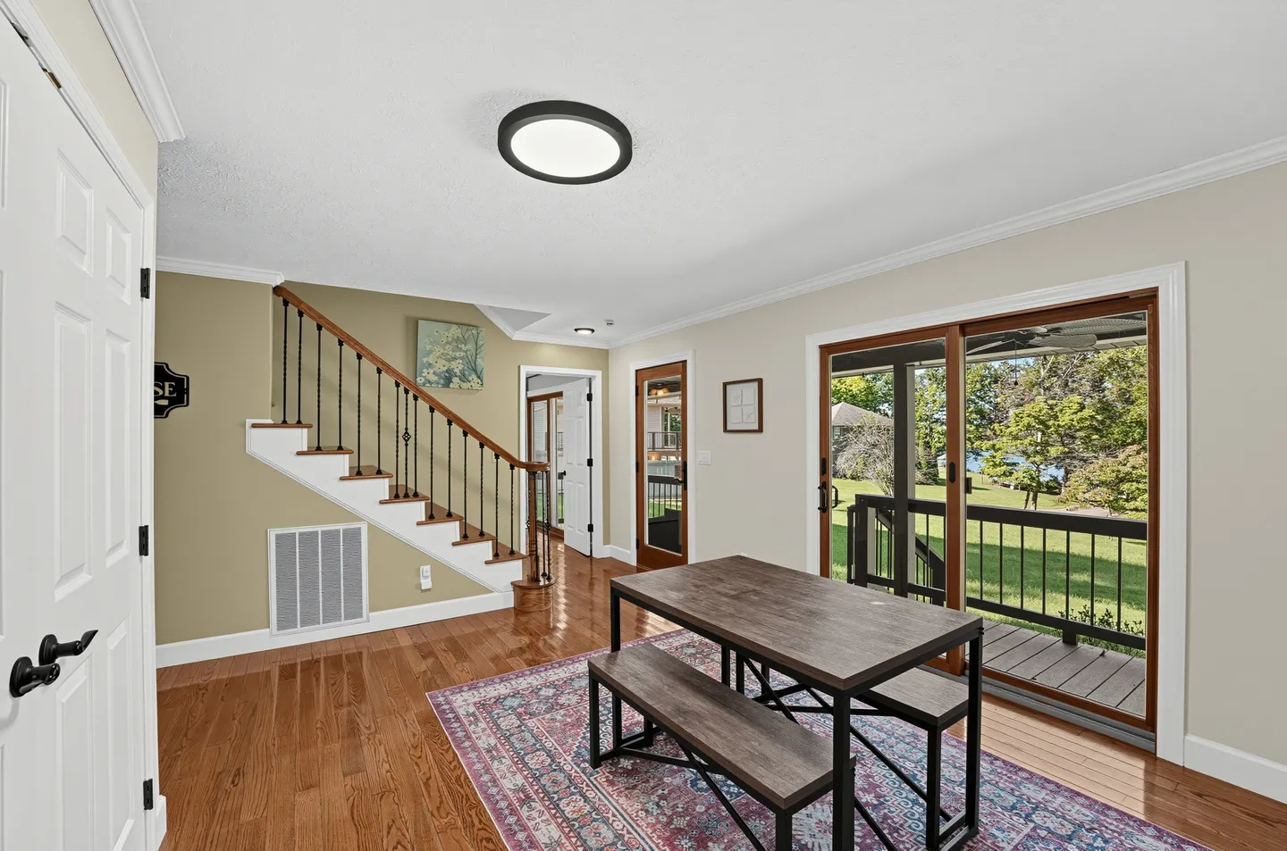 Dining area with a wood-top table and benches on a patterned rug, near a staircase and sliding glass doors leading to an outdoor deck.