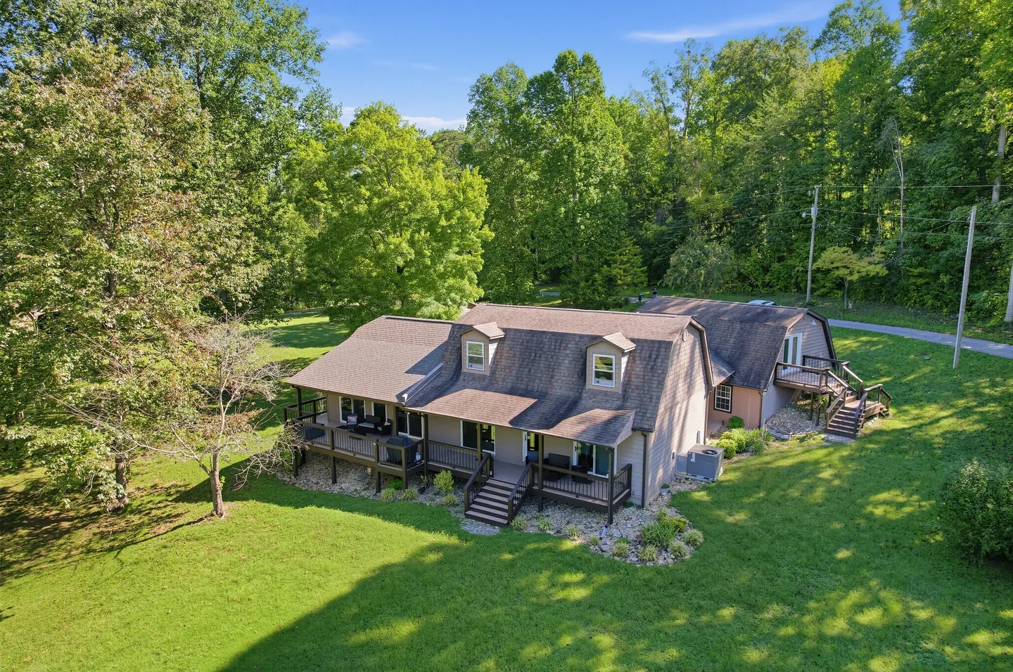 Aerial view of a two-story home with a wrap-around wooden deck surrounded by a green lawn and dense trees.