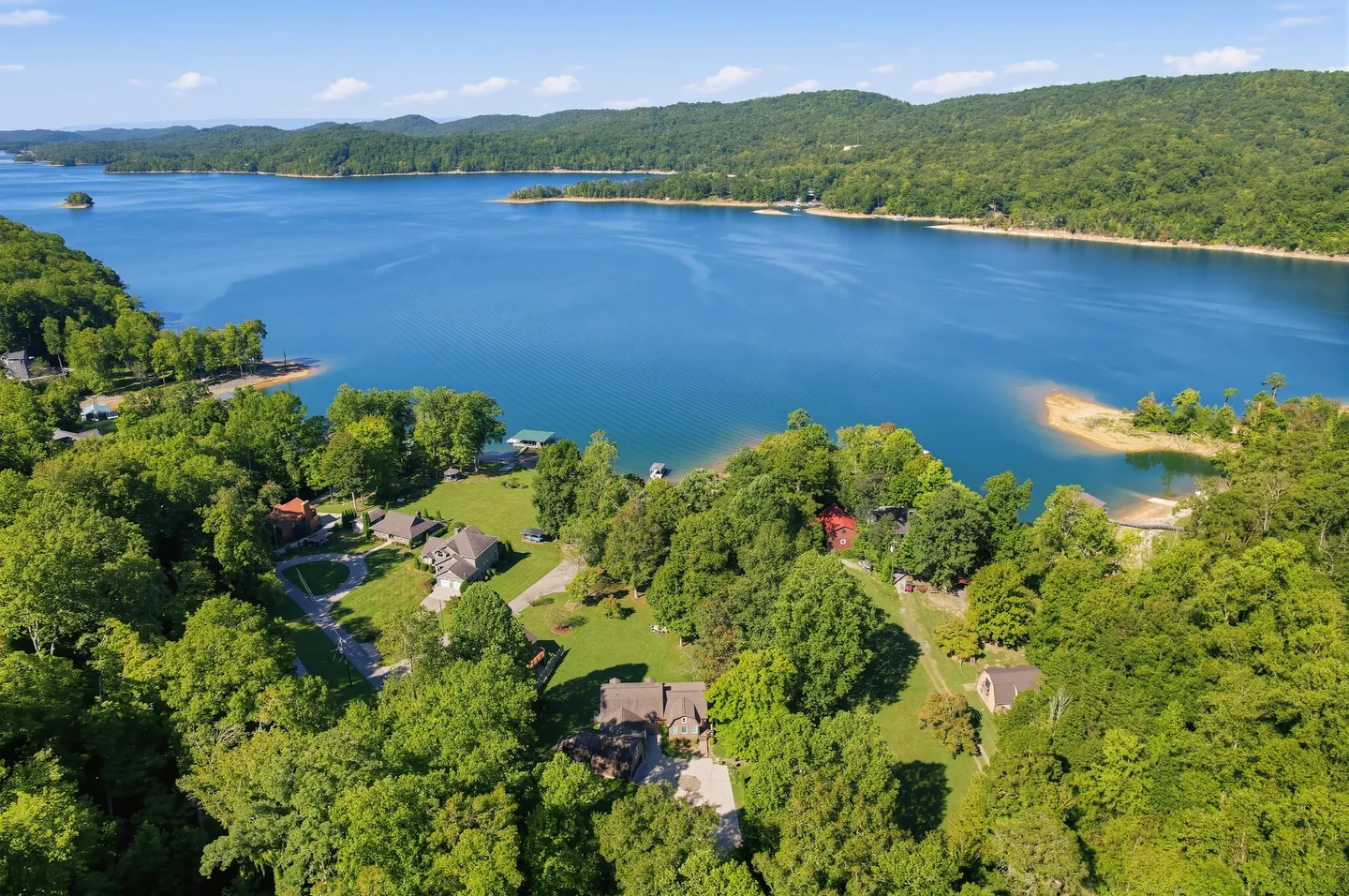 Aerial view of a home nestled among lush green trees on the shoreline of a large blue lake surrounded by forested hills under a clear sky.