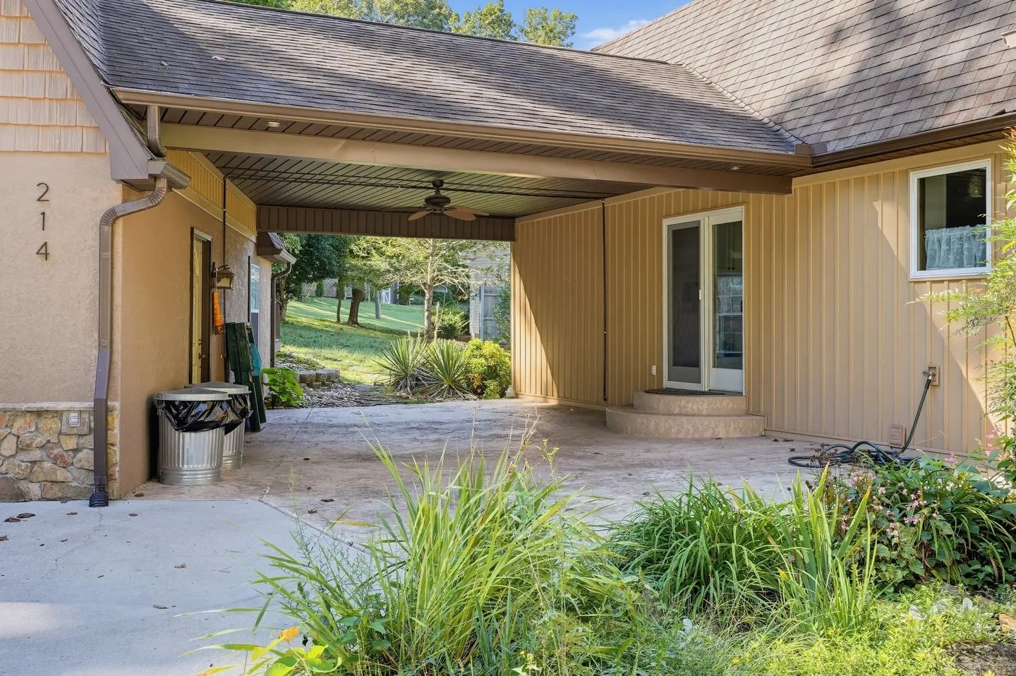 Exterior view of a covered carport or breezeway leading into a grassy backyard, adjacent to house siding and a glass patio door.