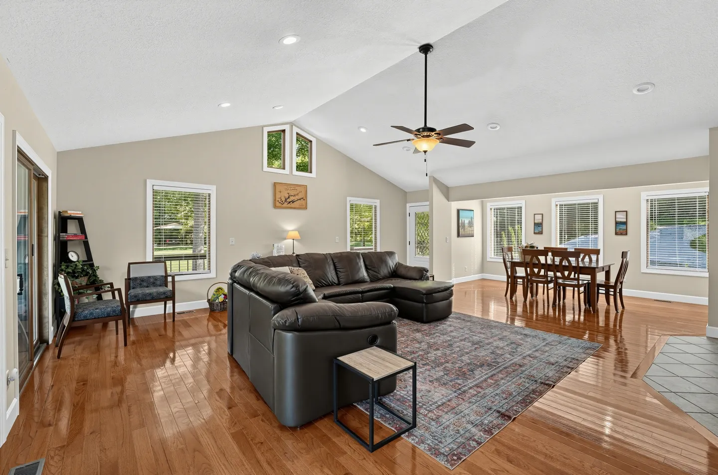 Open-concept living and dining area with vaulted ceilings, wood floors, a dark leather sectional, and a dining table.