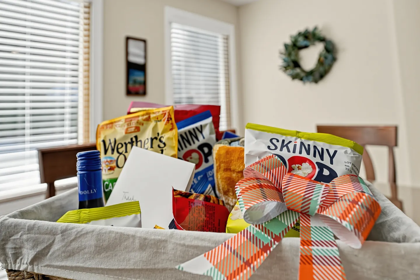 A welcome gift basket filled with assorted snacks, a card, and a bottle with a colorful plaid bow.