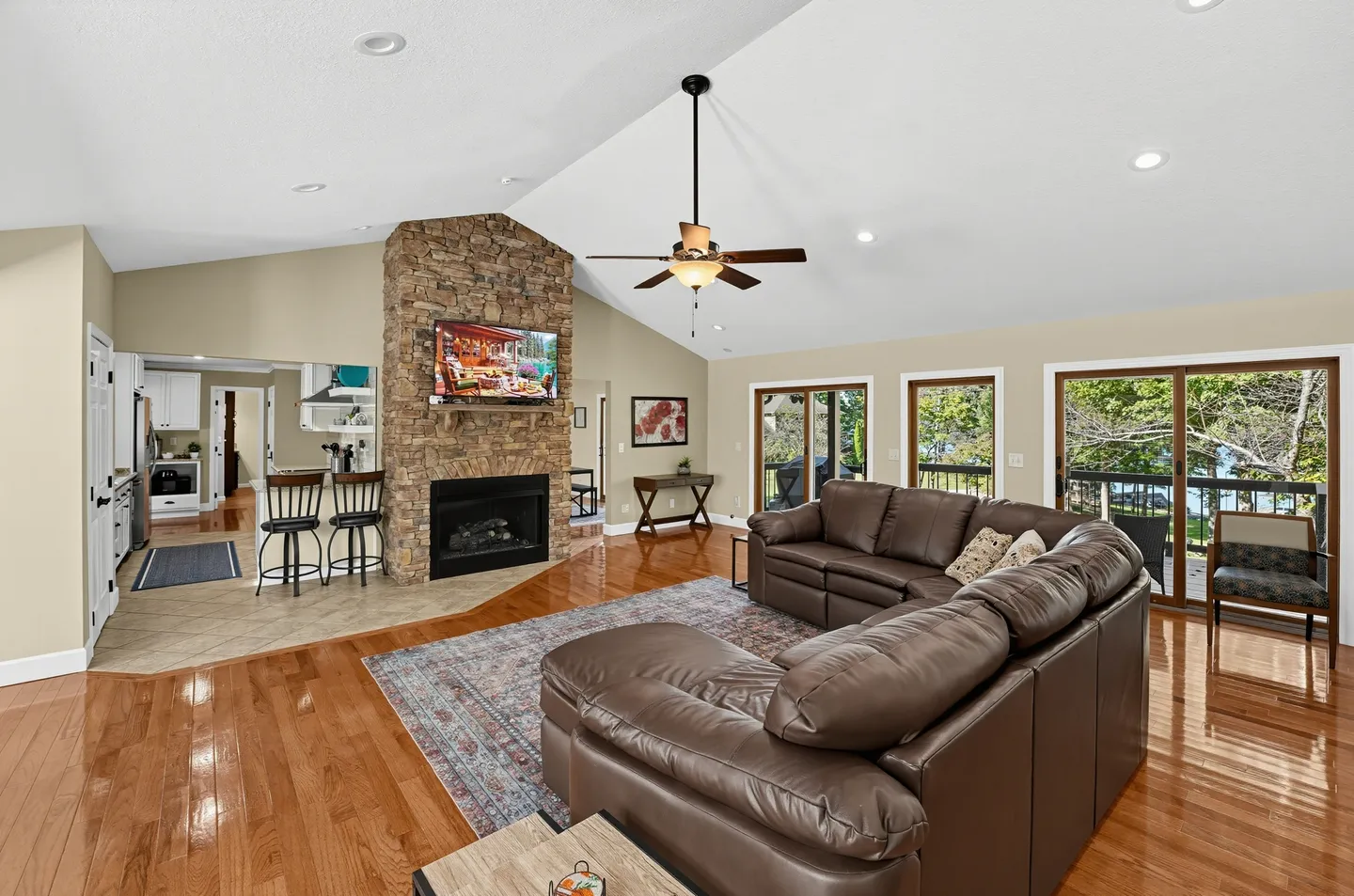 Open living area with vaulted ceilings, a leather sectional couch, a stone fireplace with a mounted television, and sliding glass doors.
