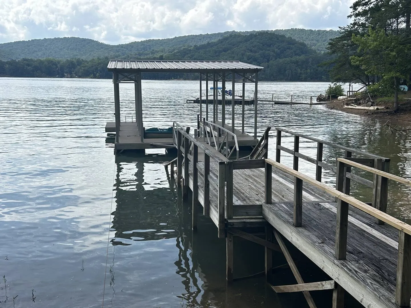 A wooden dock and gangway leading to a covered boat slip on a wide lake with forested hills in the background.