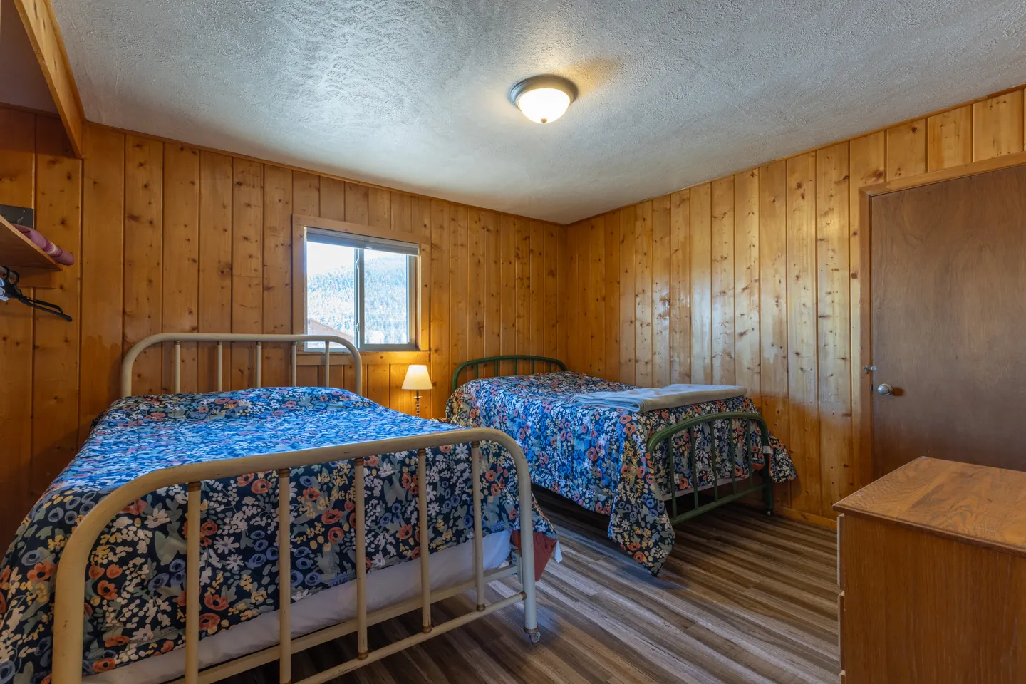 Bedroom with vertical wood paneling, two metal-frame beds with blue floral bedding, and wood-look flooring.