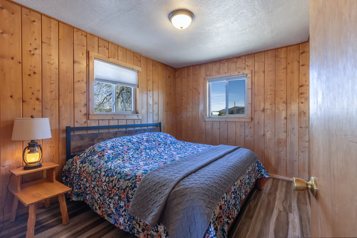Bedroom with vertical wood-paneled walls, a made bed with a blue floral spread, a small nightstand with a lamp, and two windows.