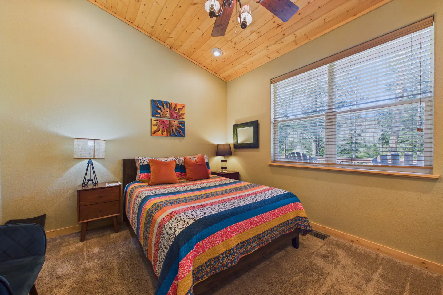 Bedroom with a patterned colorful bedspread, wood plank ceiling with ceiling fan, and large window with blinds overlooking trees.